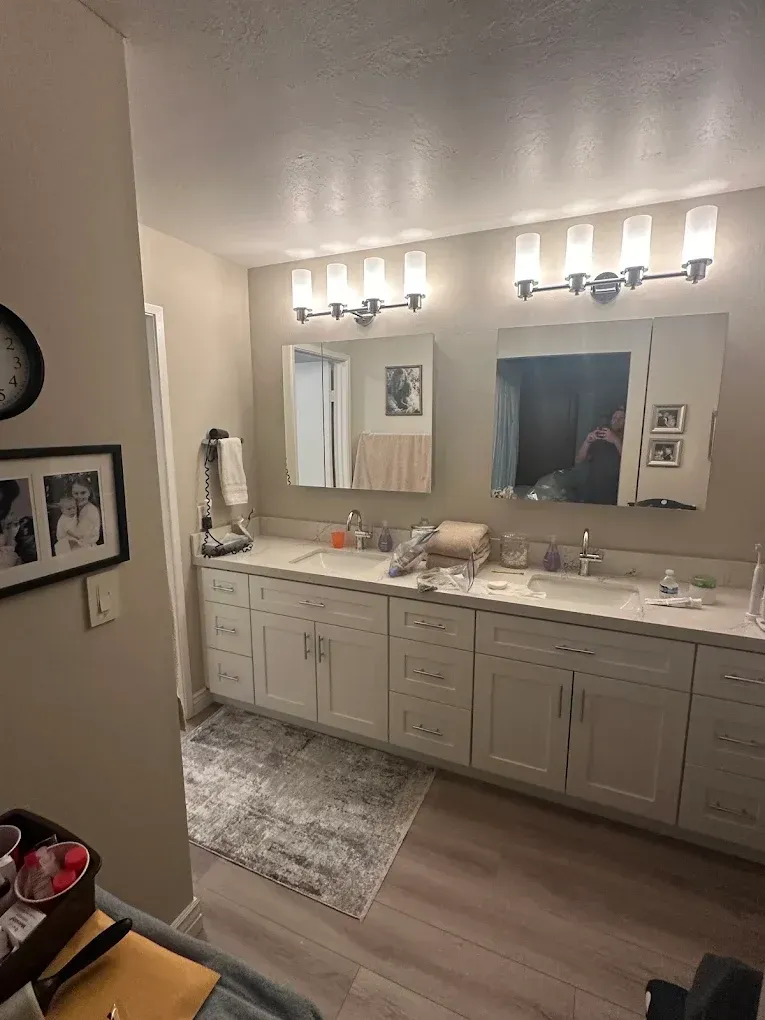 Bright bathroom vanity with large mirrors, wall lights, and white cabinetry over wood-look flooring.