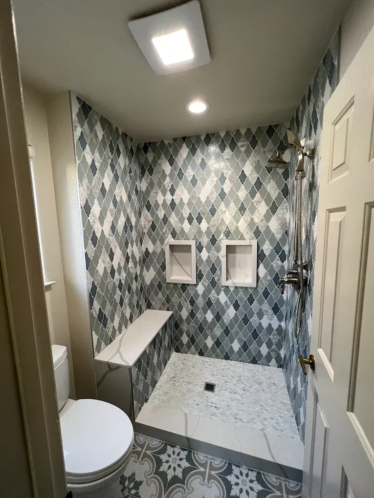 Small bathroom with patterned tile walls, shower, toilet, and a wall-mounted bench.