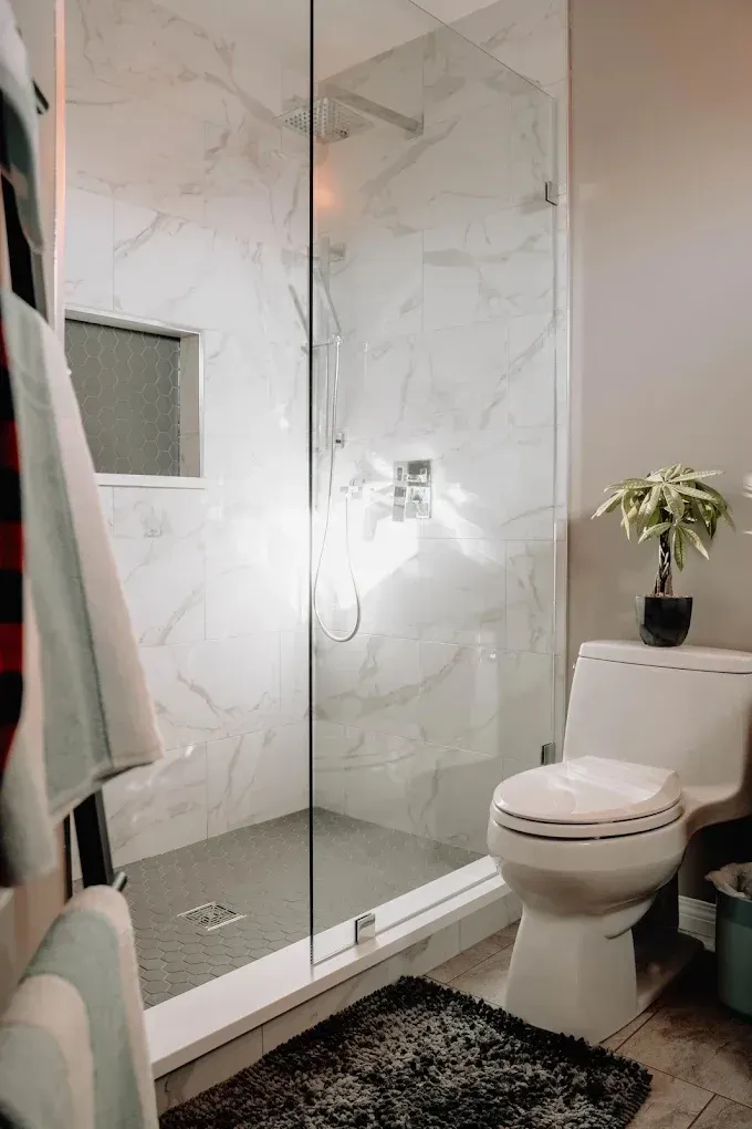 Modern bathroom with a glass shower, white marble walls, toilet, and small potted plant