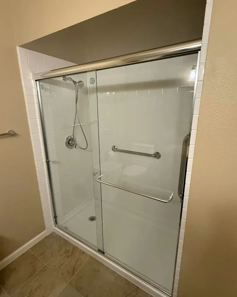 Walk-in shower with clear glass door, chrome grab bar, and beige tile walls.