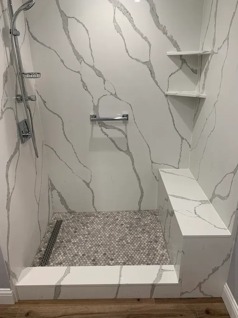 Modern walk-in shower with white marble-look tile, handheld shower, corner shelves, and pebble floor.