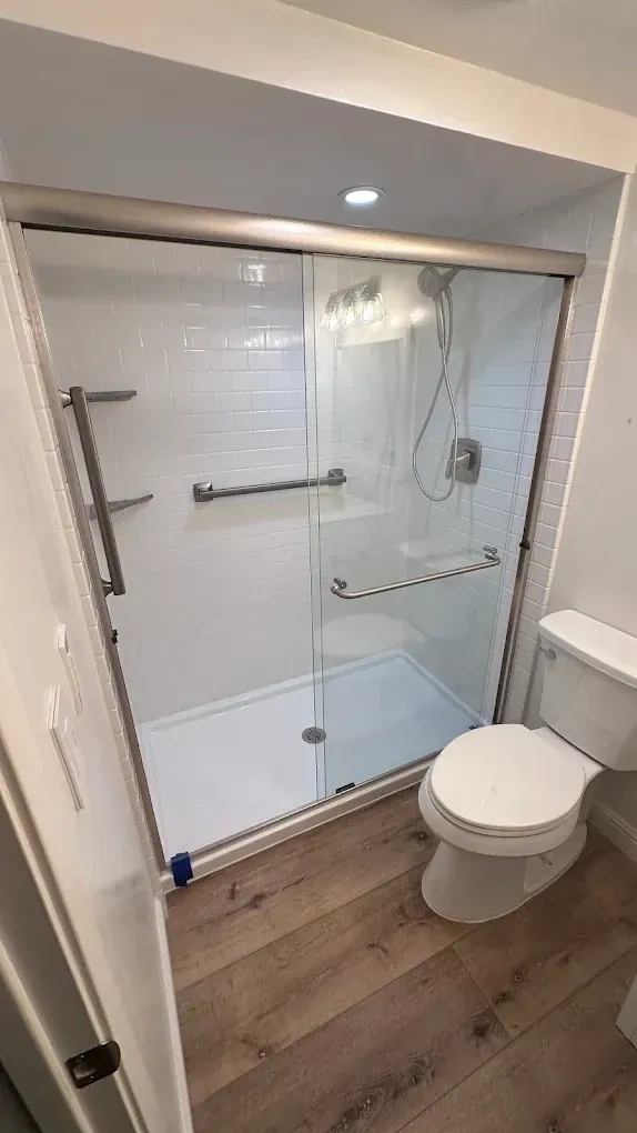 Accessible bathroom with glass walk-in shower, grab bars, and toilet on wood-look floor.