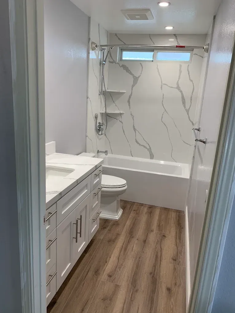 Modern bathroom with white vanity, toilet, tub-shower, and wood-look floor, seen through a doorway