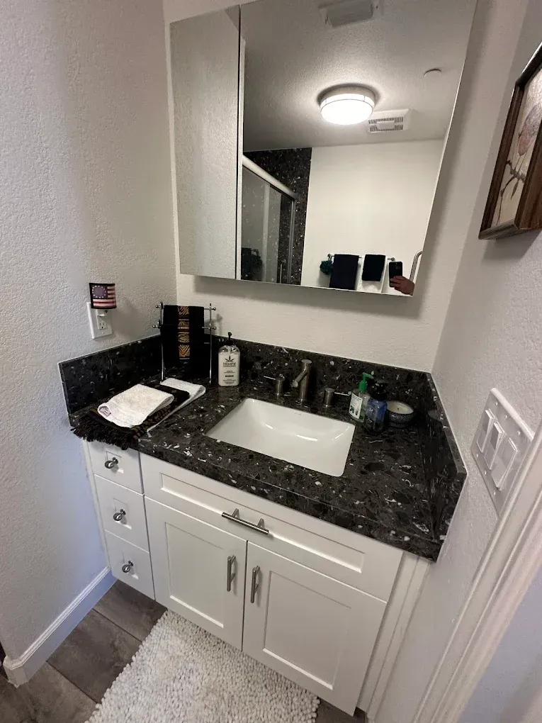 Small bathroom vanity with black granite counter, white cabinets, sink, mirror, and overhead light.