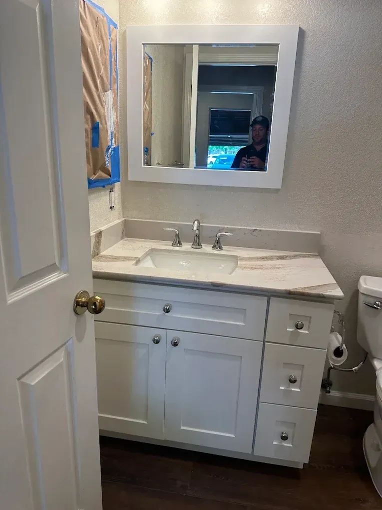 Small bathroom vanity with white cabinets, marble countertop, sink, mirror, and toilet visible on the right.