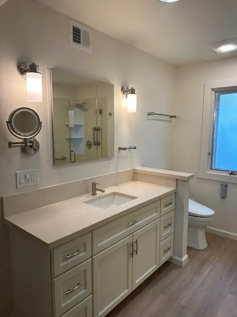 Bright bathroom with double vanity, mirror, wall lights, towel bar, and window beside a toilet.
