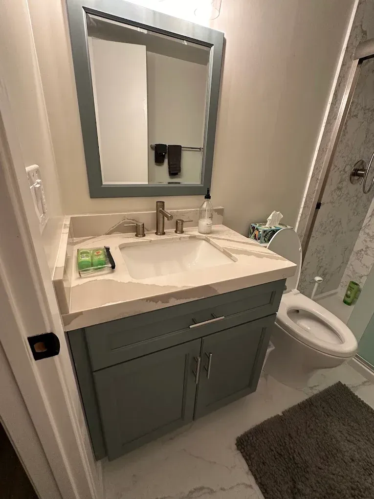 Small bathroom vanity with sink, mirror, and toilet beside a gray bath mat.
