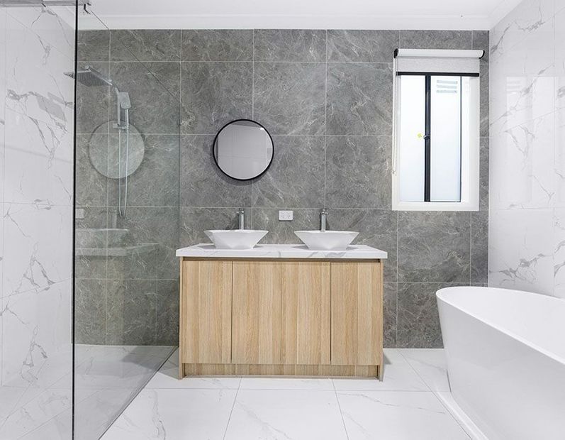 Modern bathroom with double vanity, gray tile walls, round mirror, and glass shower enclosure