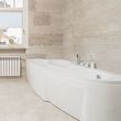 Modern bathroom with a large white bathtub, beige stone walls, window, and radiator
