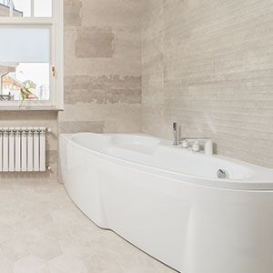 Modern bathroom with a large white bathtub, beige stone walls, window, and radiator