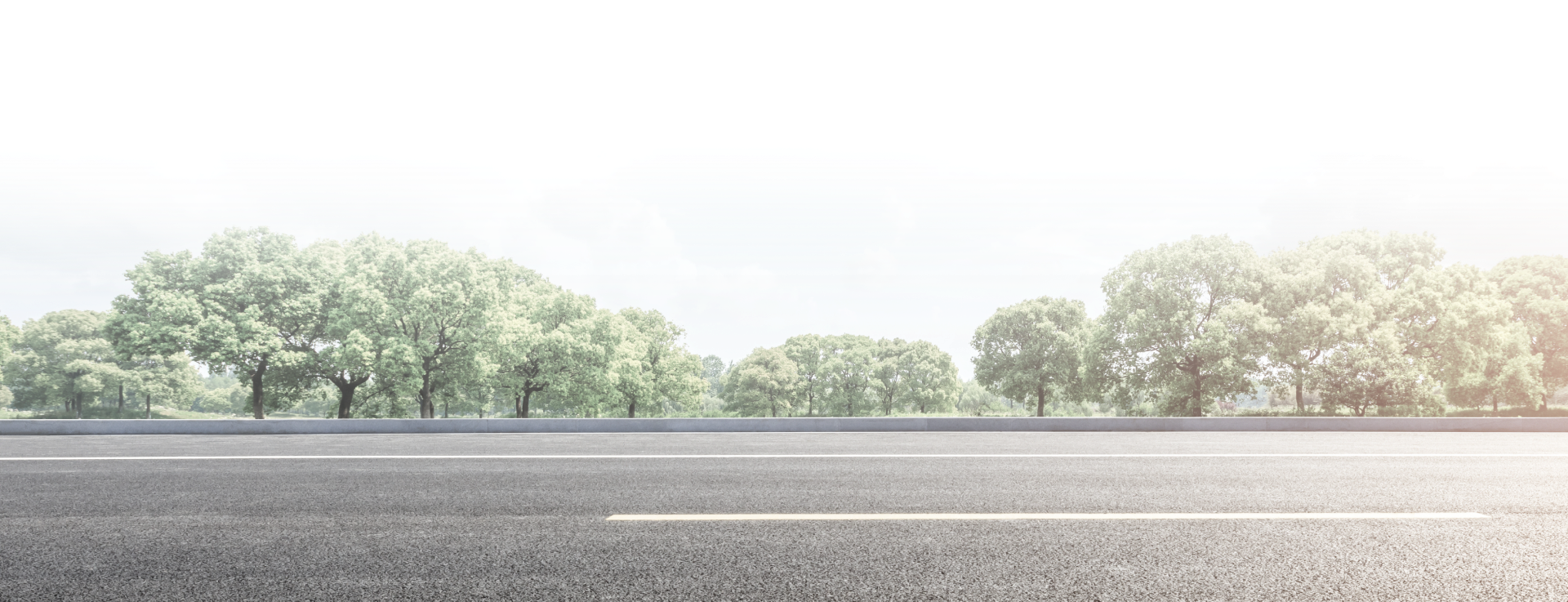 An empty asphalt road stretches horizontally across the foreground, with a line of lush green trees under a bright sky. | Avon Auto & Truck