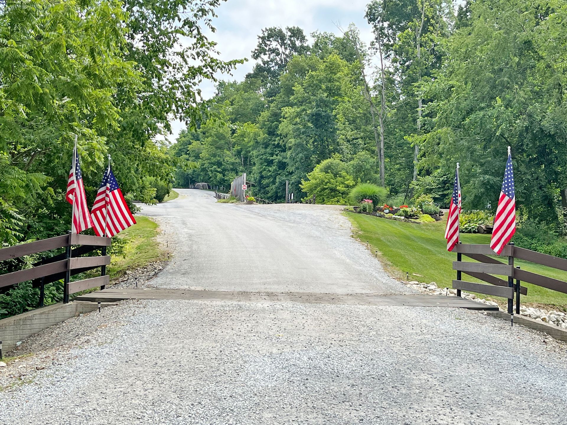 Gravel driveway with American flags, wooden fence, and trees.