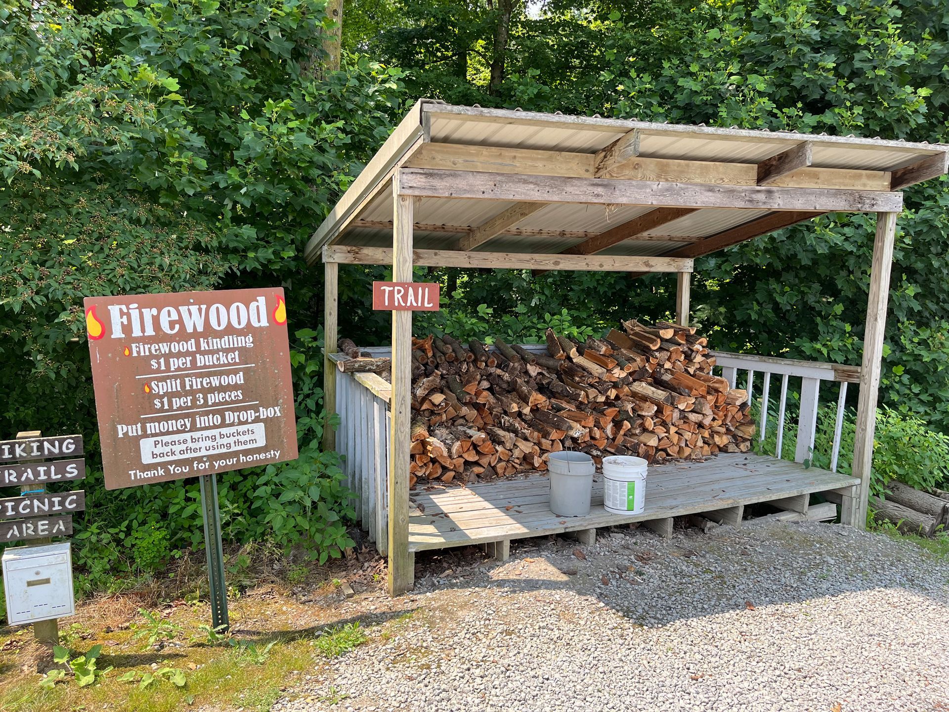Wood shed with firewood, sign, and trail markers in a wooded area.