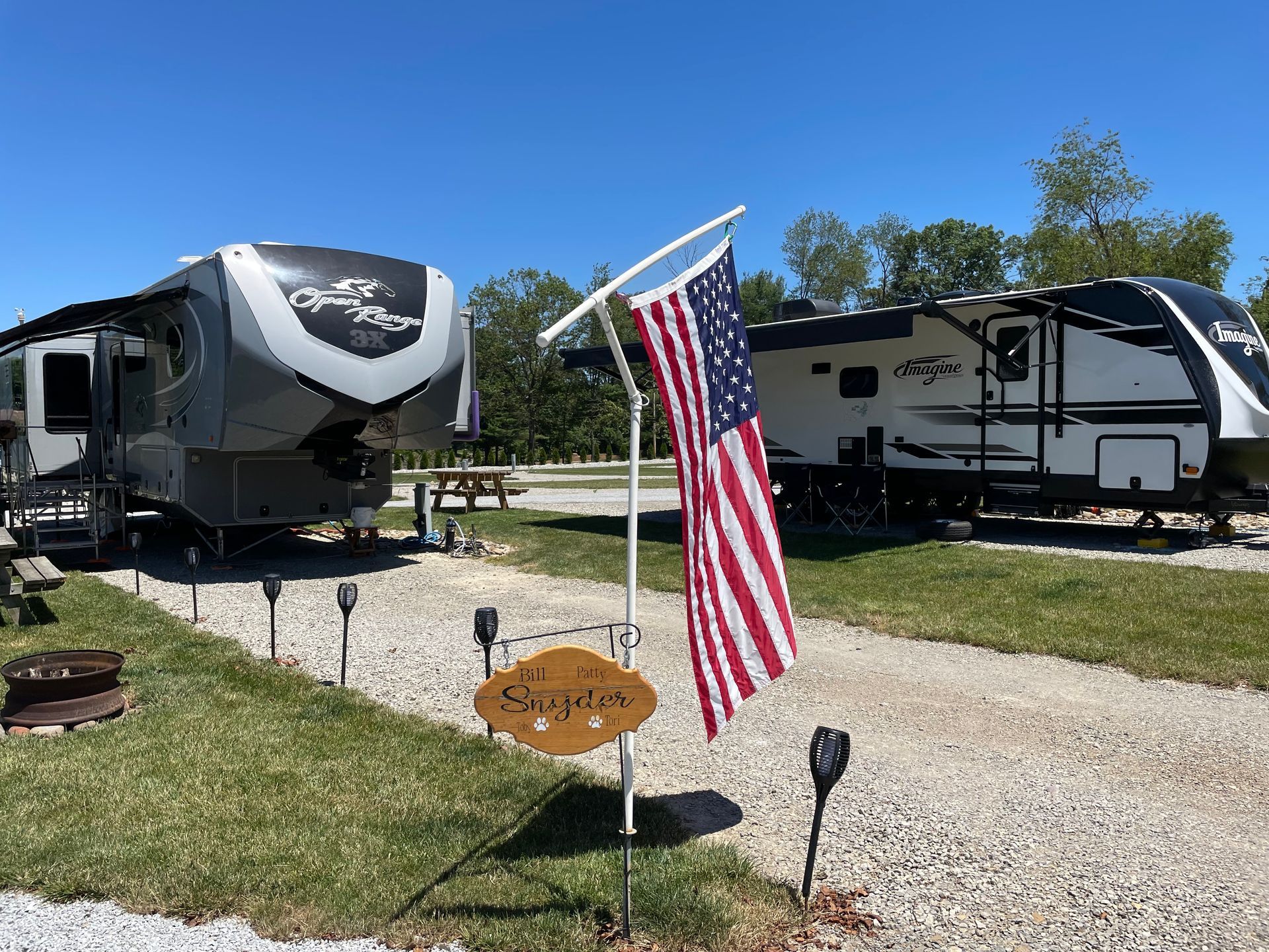 Two RVs parked in a campground; American flag, gravel drive, green grass, and solar lights.