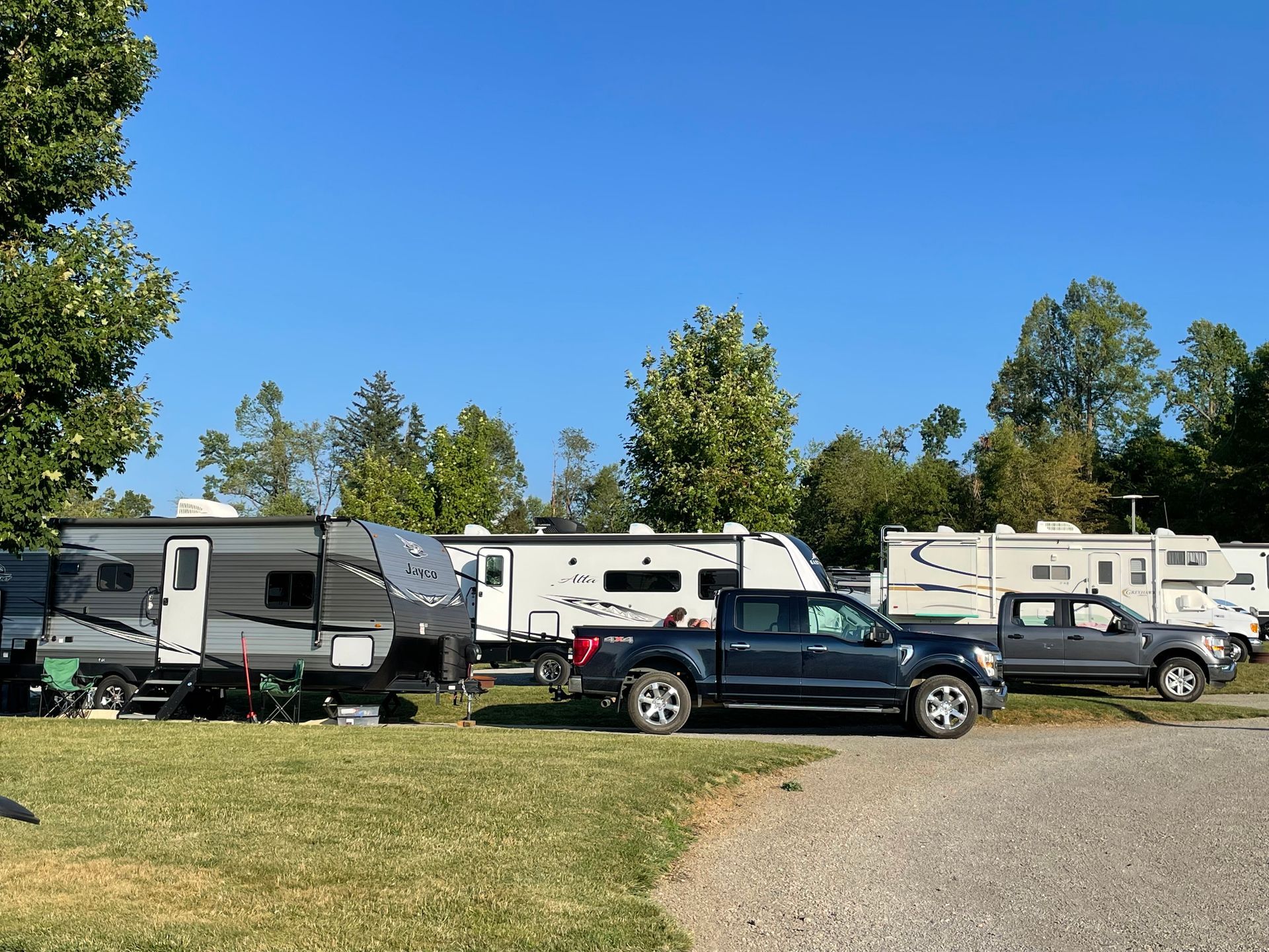 Campground with RVs parked in a row, sunny day, blue sky, trees in the background.