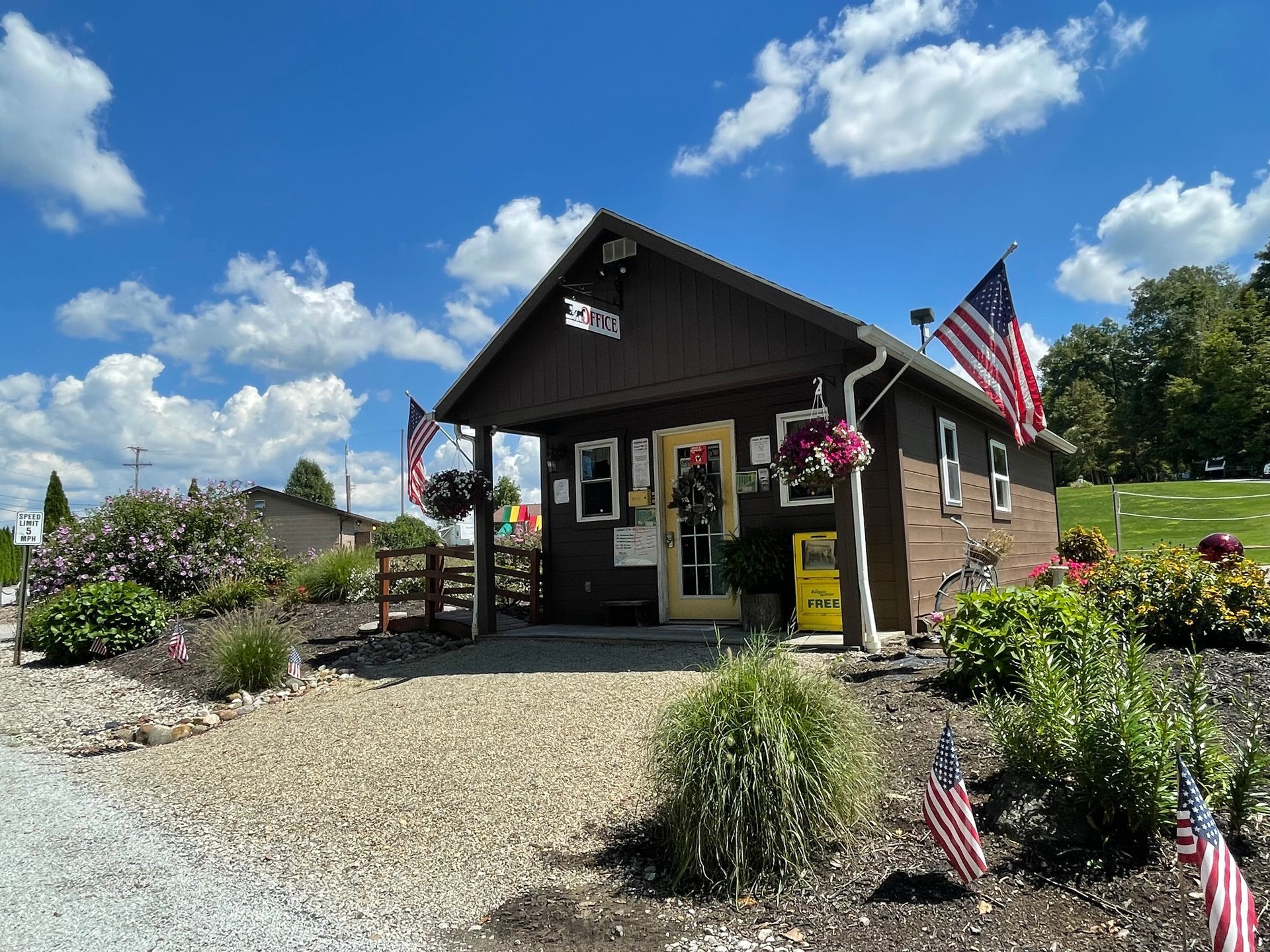 Brown building with US flags and flower baskets. Gravel path, blue sky.
