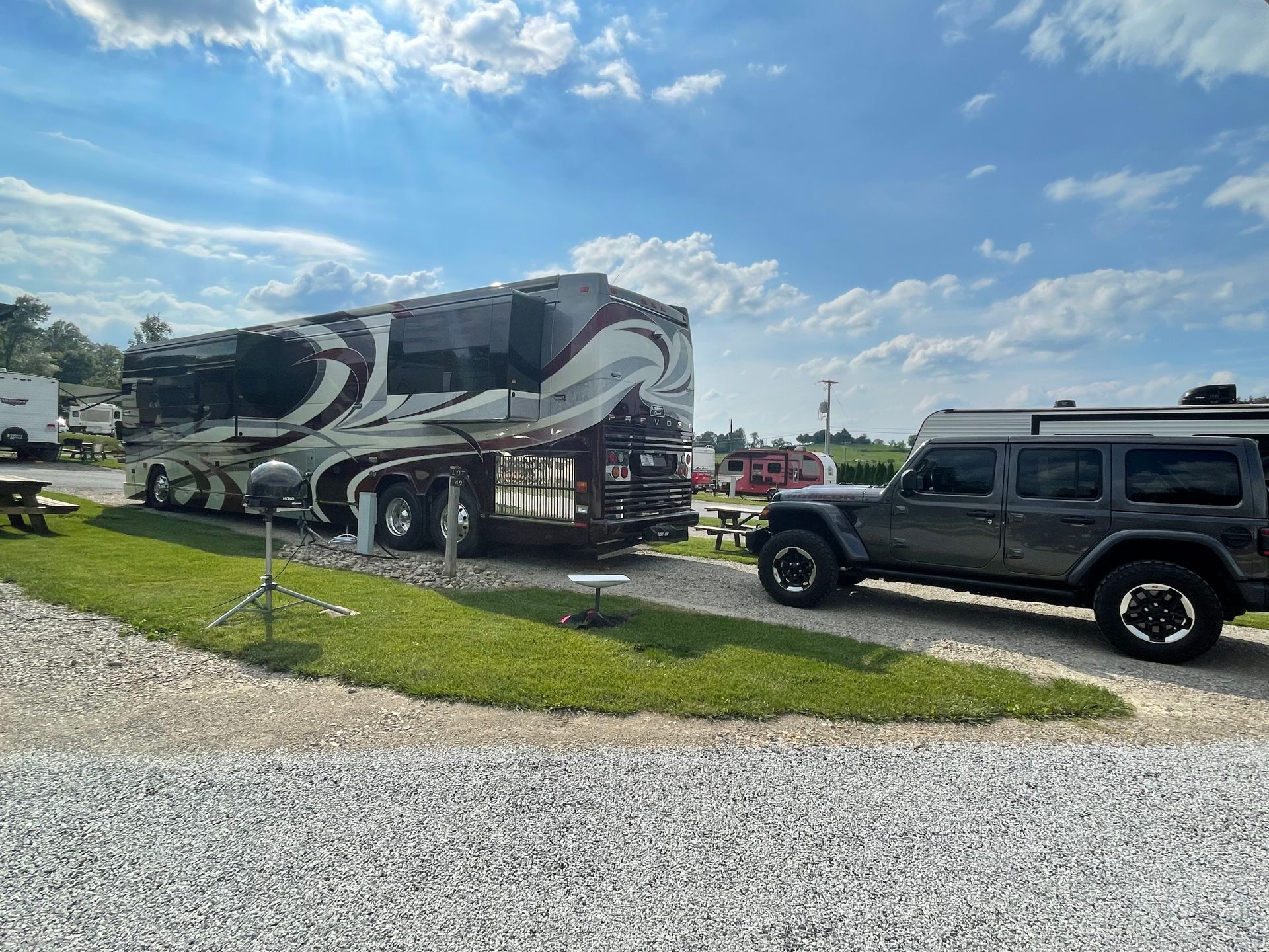 Large RV and gray Jeep parked on gravel at a campground under a blue sky.