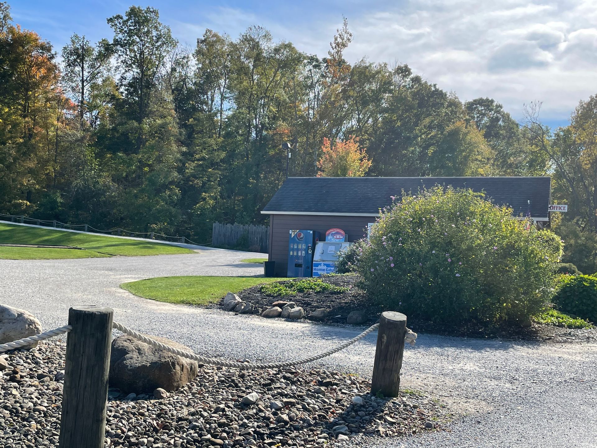 Gravel driveway leads to a small building surrounded by trees. A chain fence and posts are in the foreground.