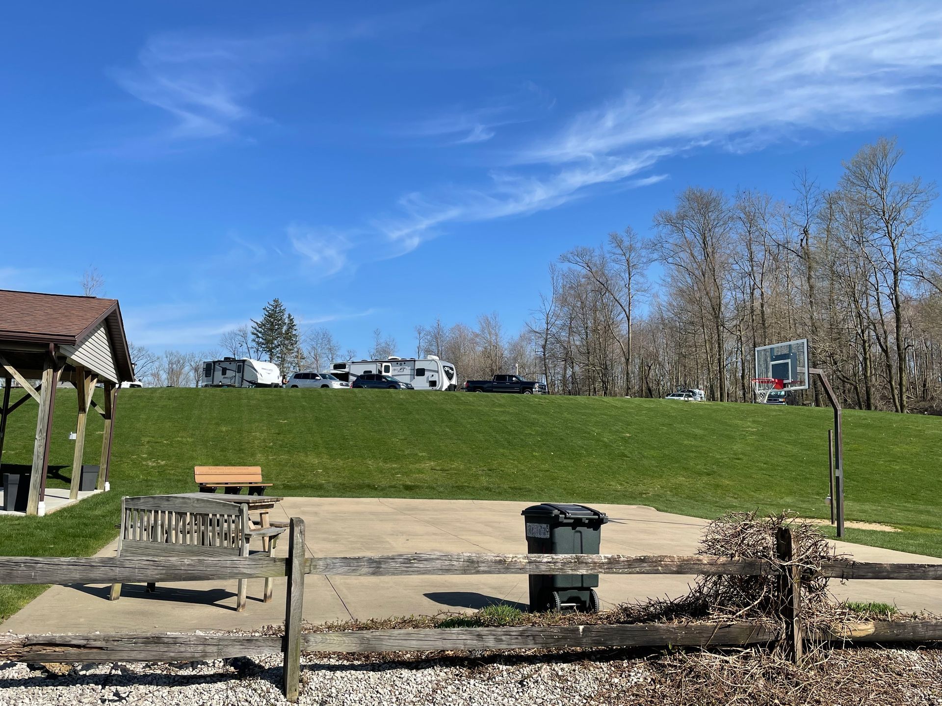 A park scene with a basketball court, picnic area, and RVs on a grassy hill under a blue sky.