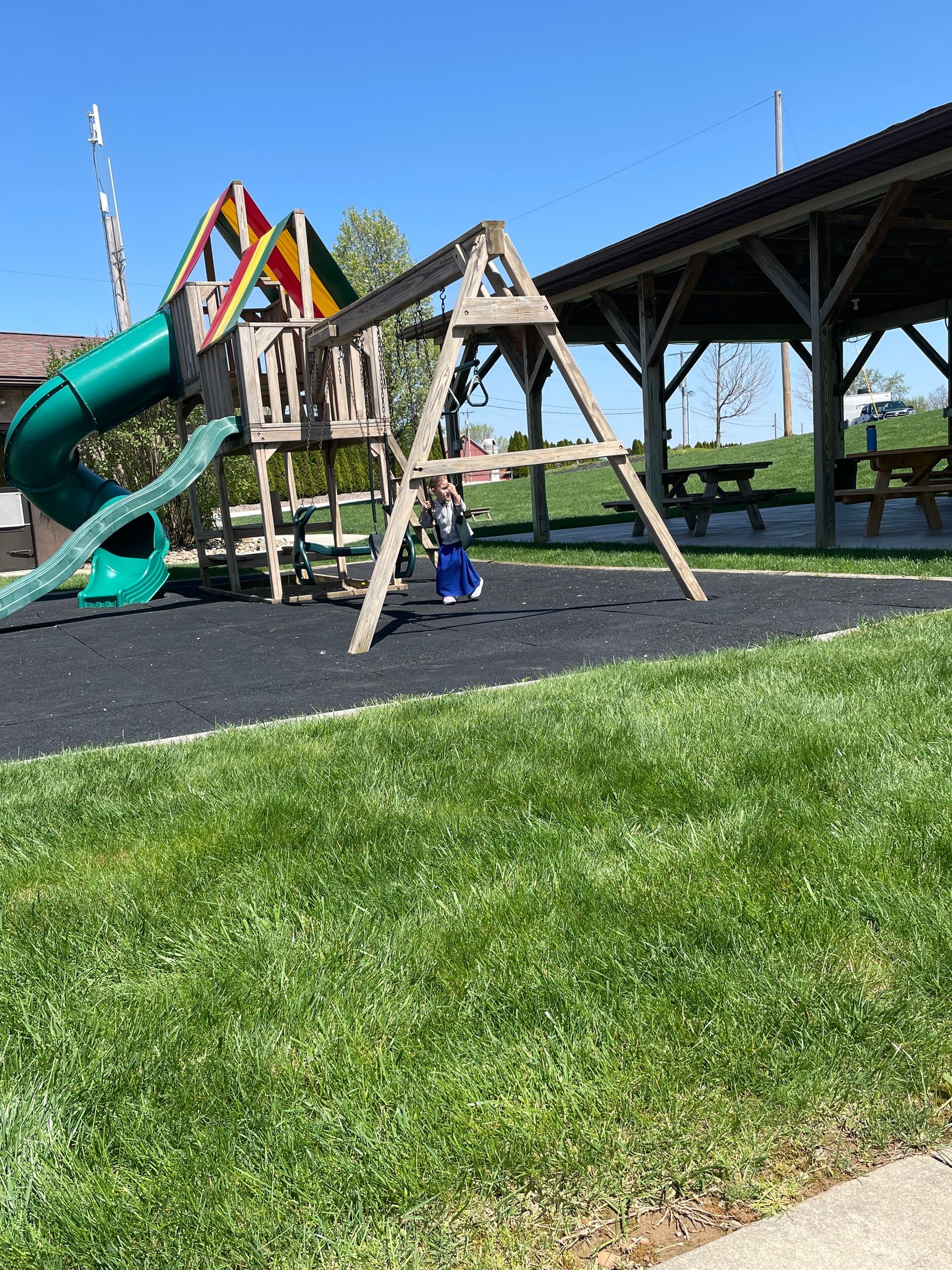 Playground with a swing set and slide, next to a covered picnic area. Green grass in the foreground.