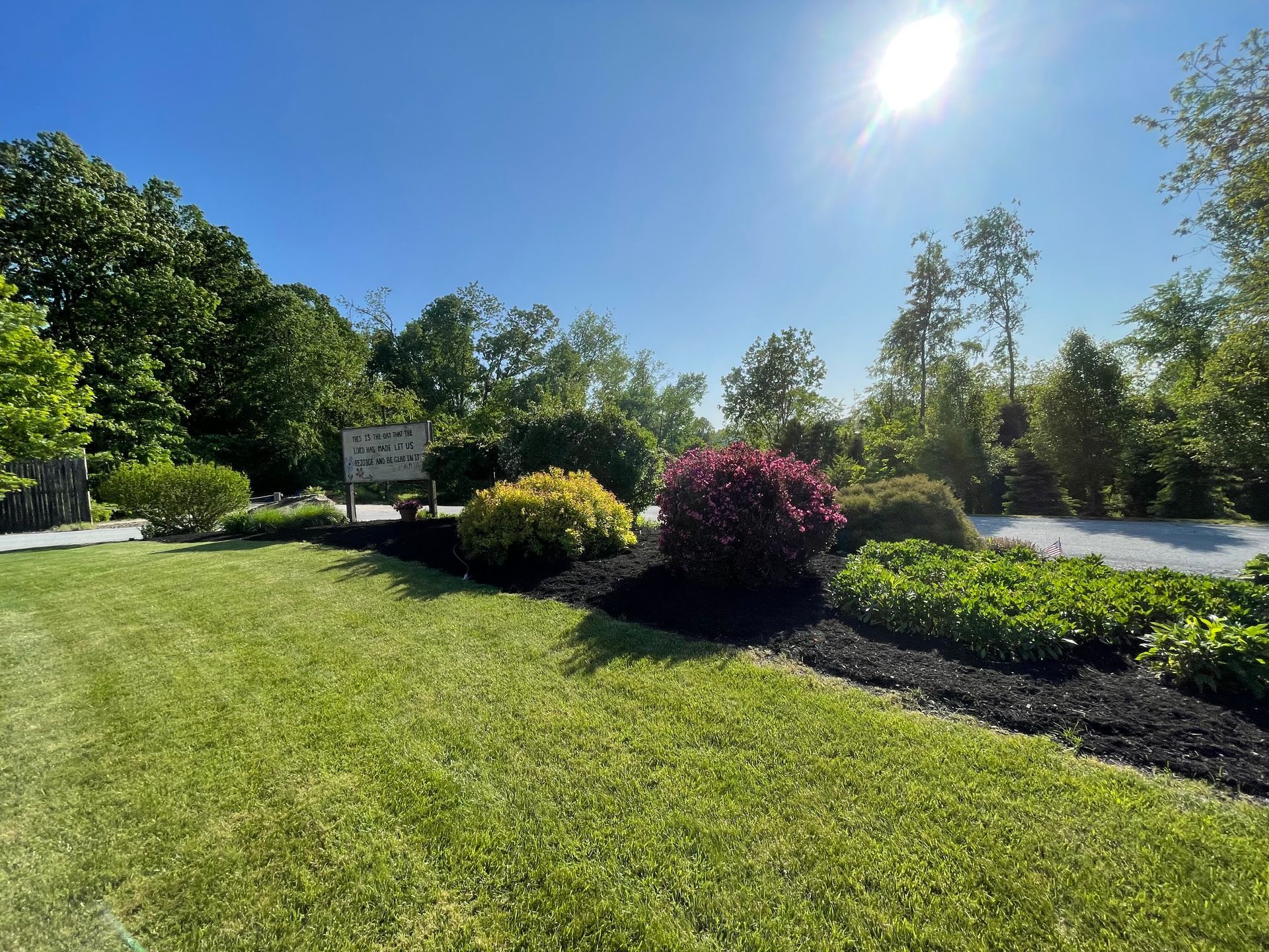 Lush green lawn with a bed of flowers in front of a sign and trees under a sunny sky.