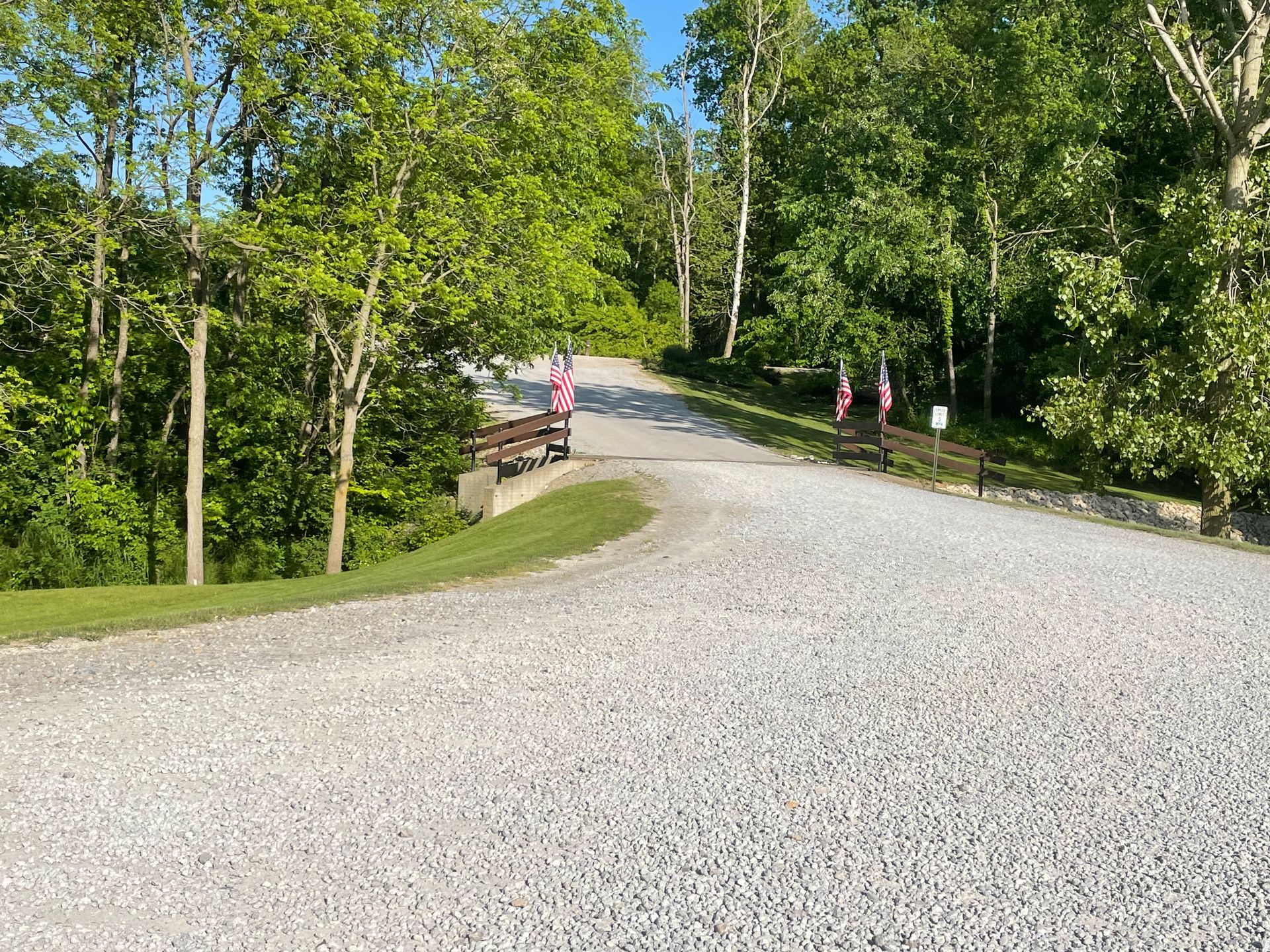 Gravel road leading uphill, bridge with flags, surrounded by trees and greenery under a sunny sky.
