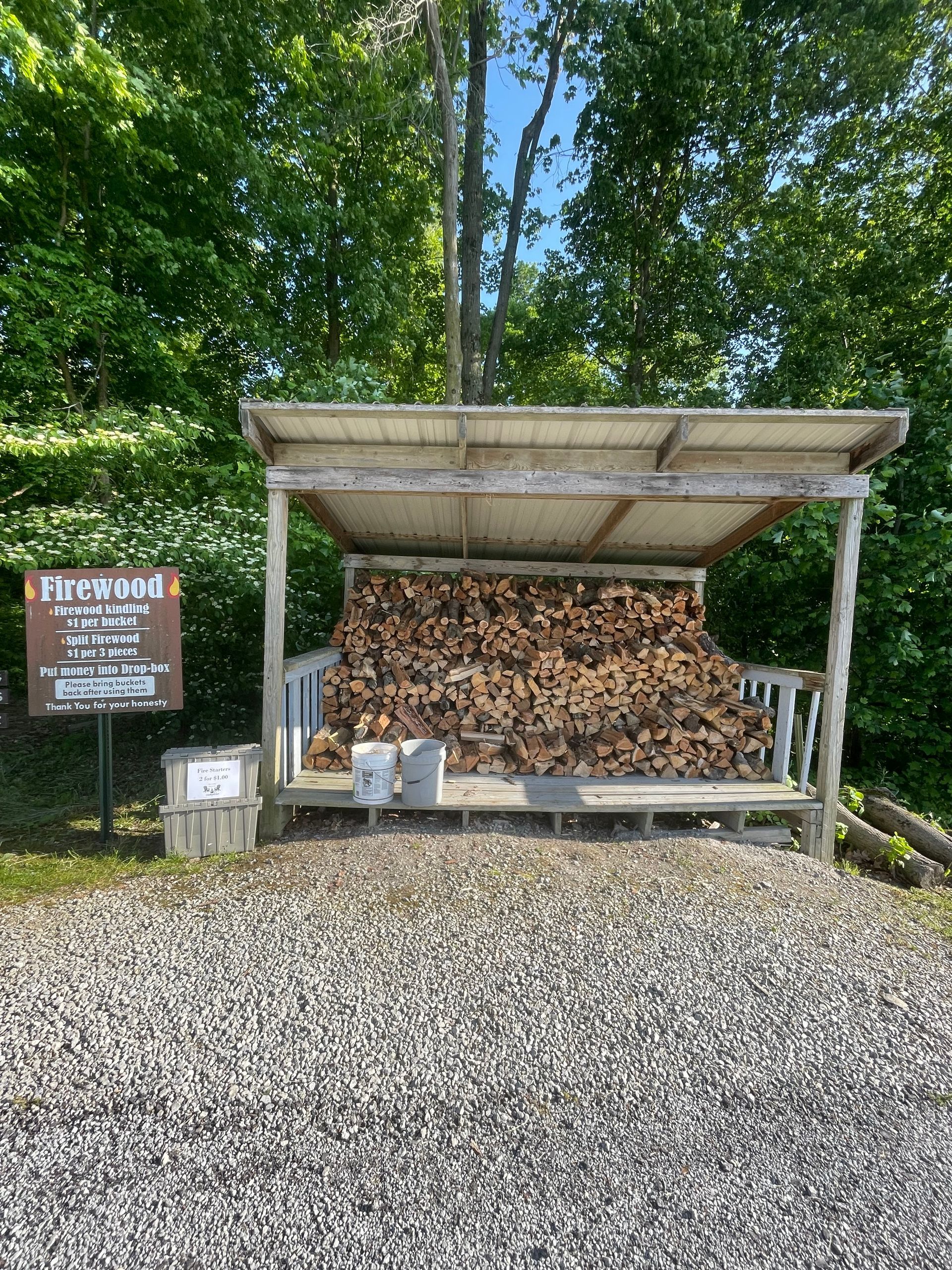 Pile of firewood stacked under a wooden shelter, gravel ground, sign on the left.