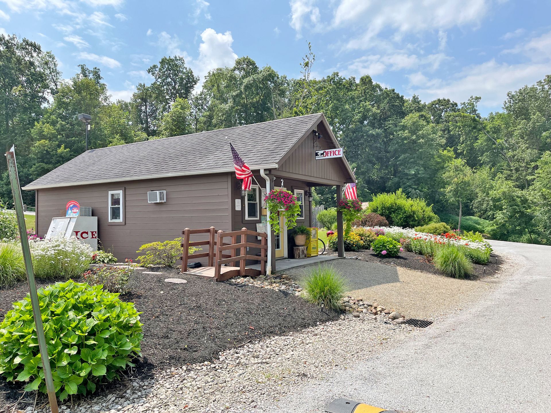 Small brown building with flowers and American flags; gravel drive, trees in background.