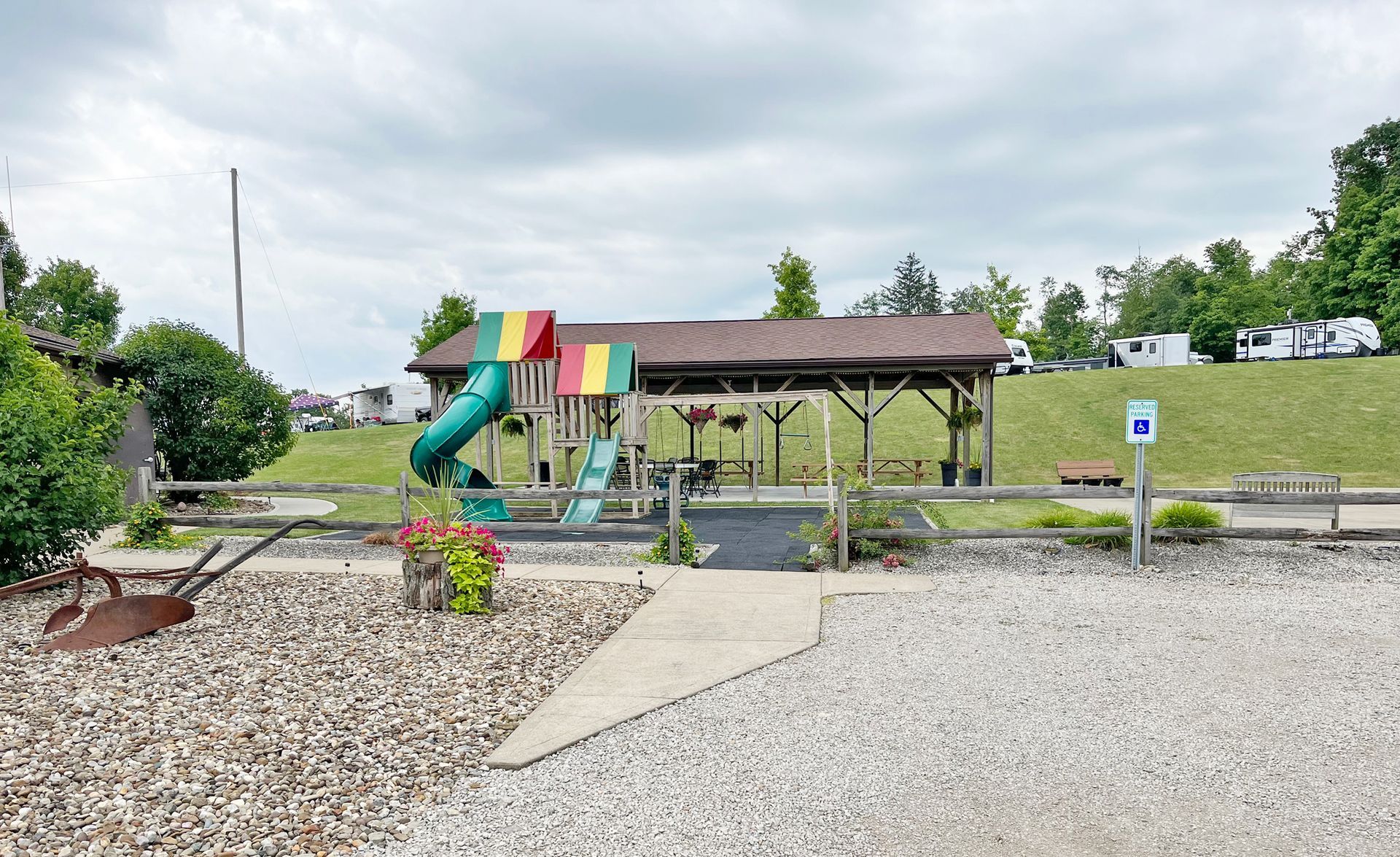Playground with slide and covered picnic area on a gravel area with green vegetation and cloudy sky.