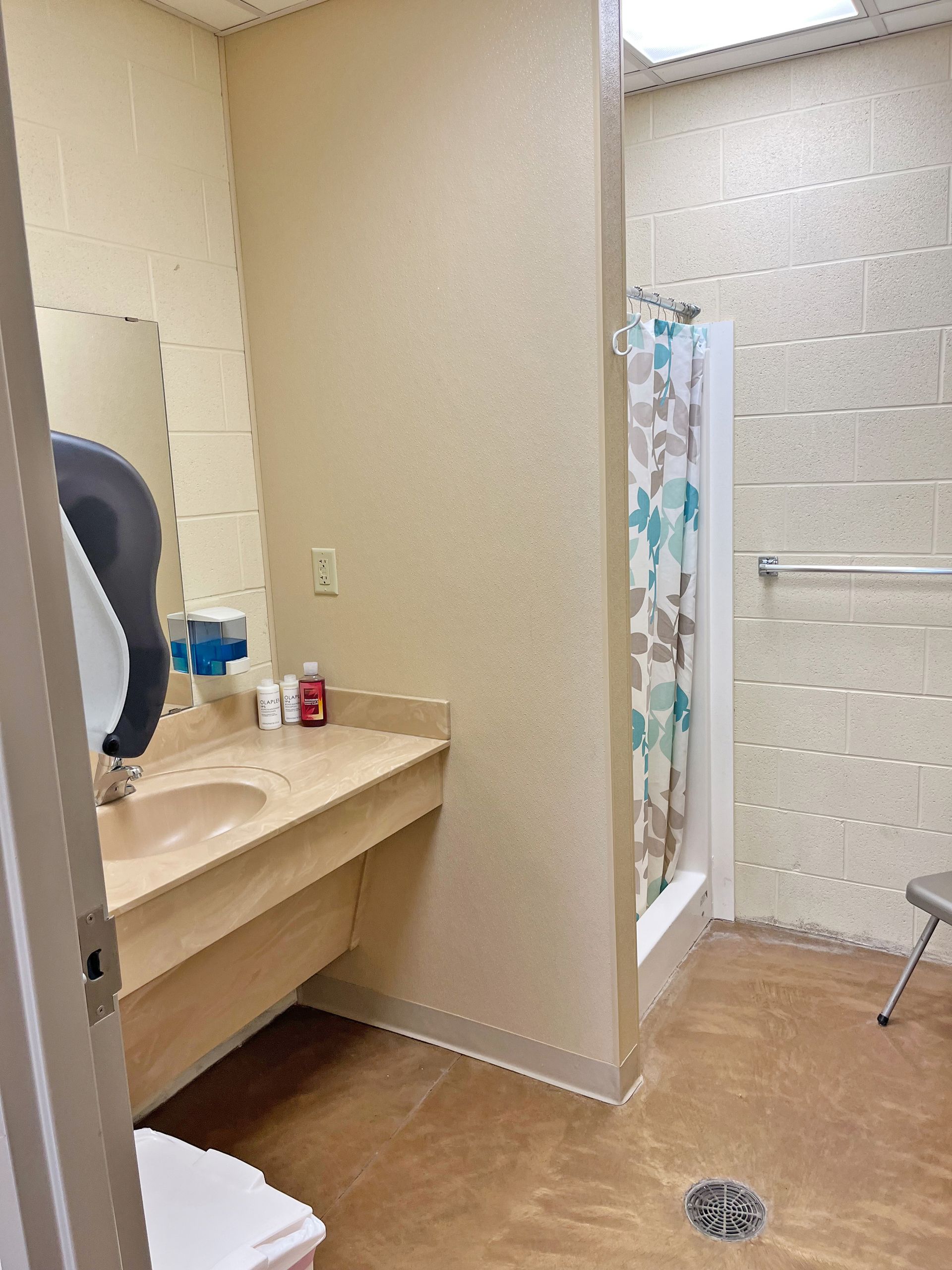 Bathroom with a sink, mirror, and shower with a floral curtain. Brown flooring and tan walls.