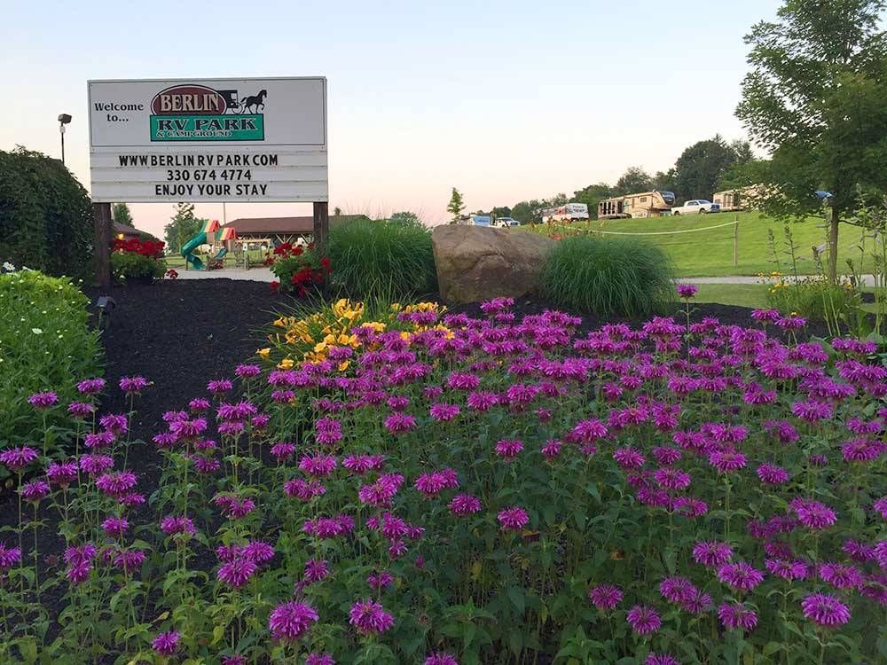 Sign for Berle's Campground in front of purple flowers, with green bushes and a grassy hill in the background.
