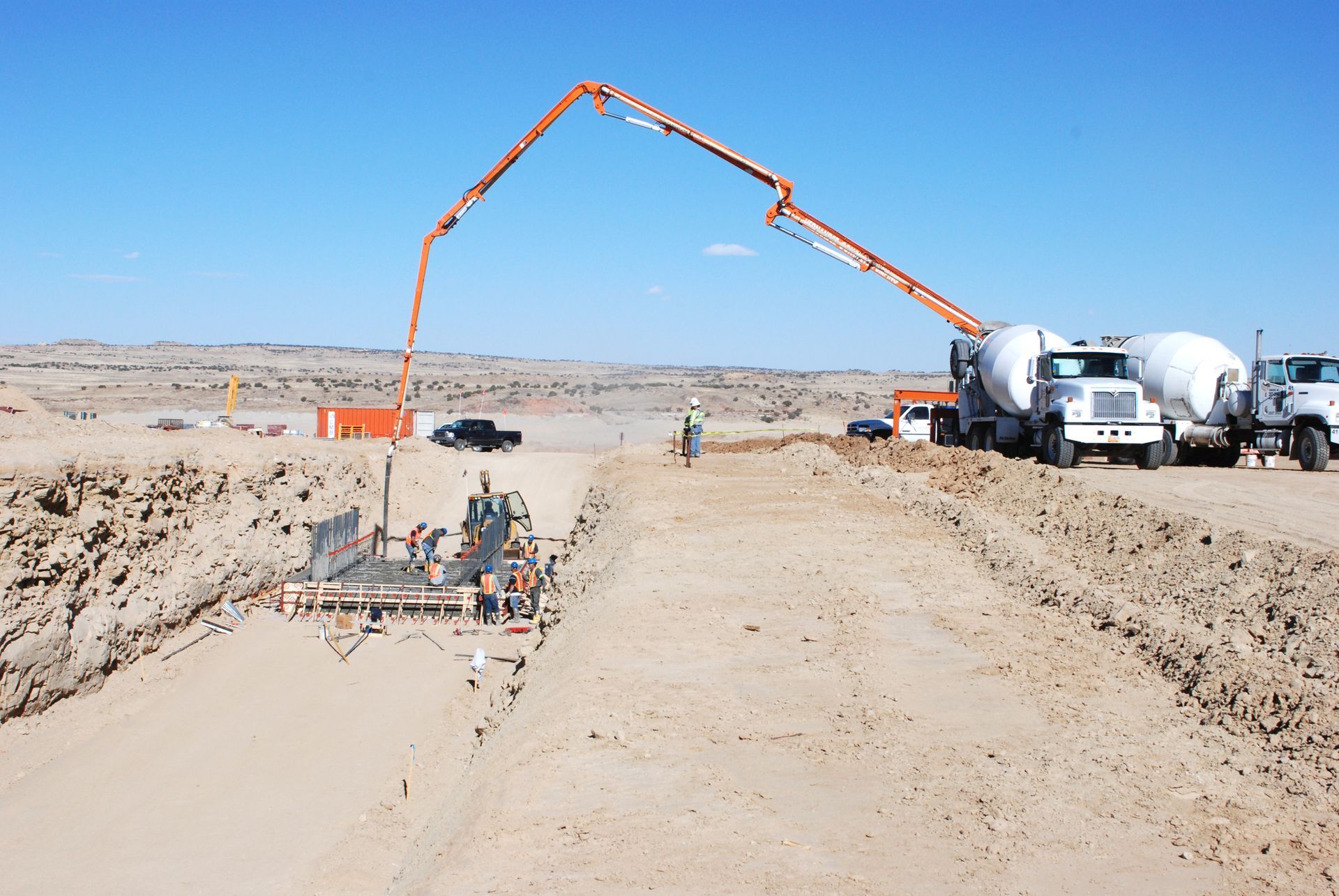 Concrete pump pouring at a road construction site with trucks and workers on a dusty desert road