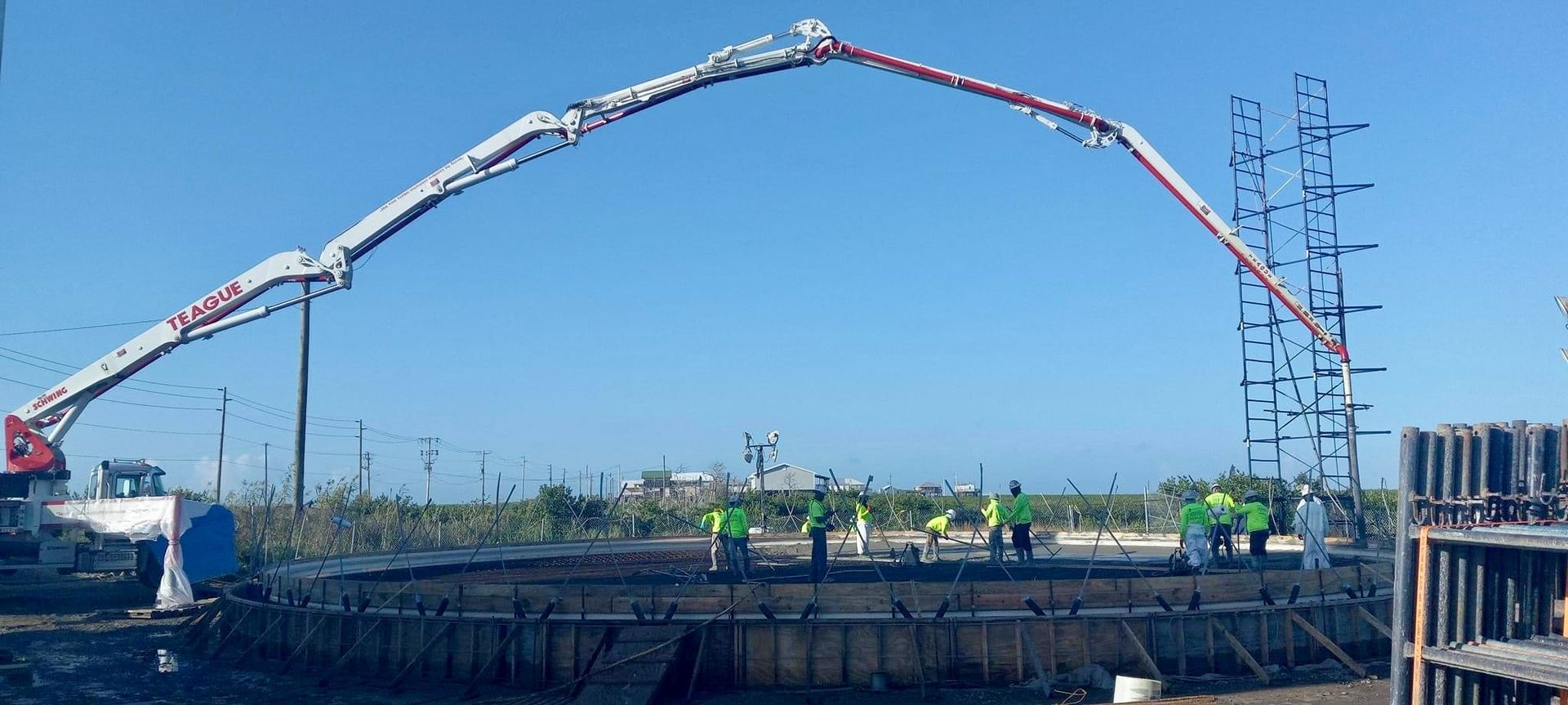 Concrete pump pouring concrete into a round foundation; construction workers in neon vests. Blue sky.