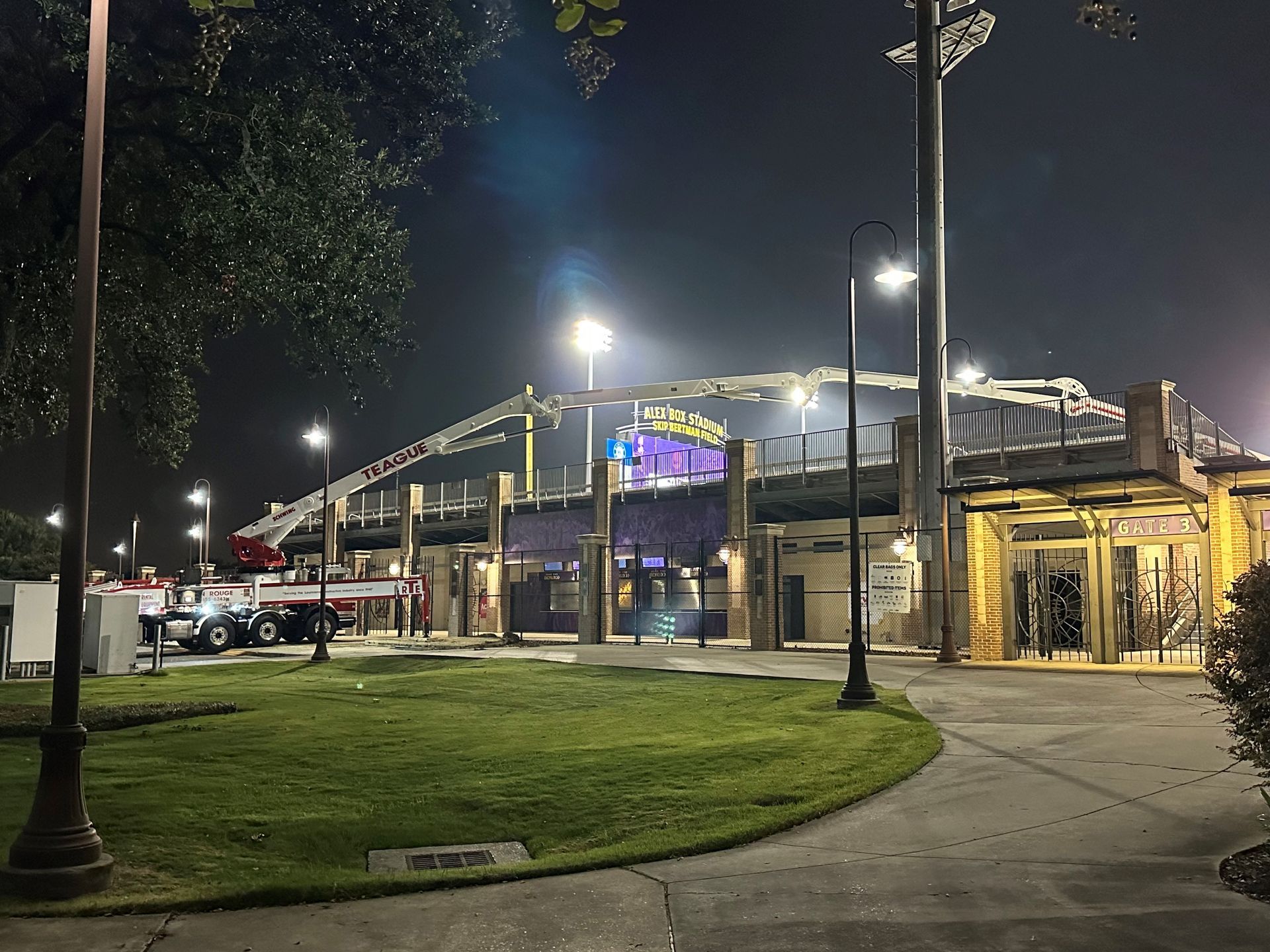 Stadium exterior at night, lit by spotlights. Construction cranes visible, green lawn in foreground.