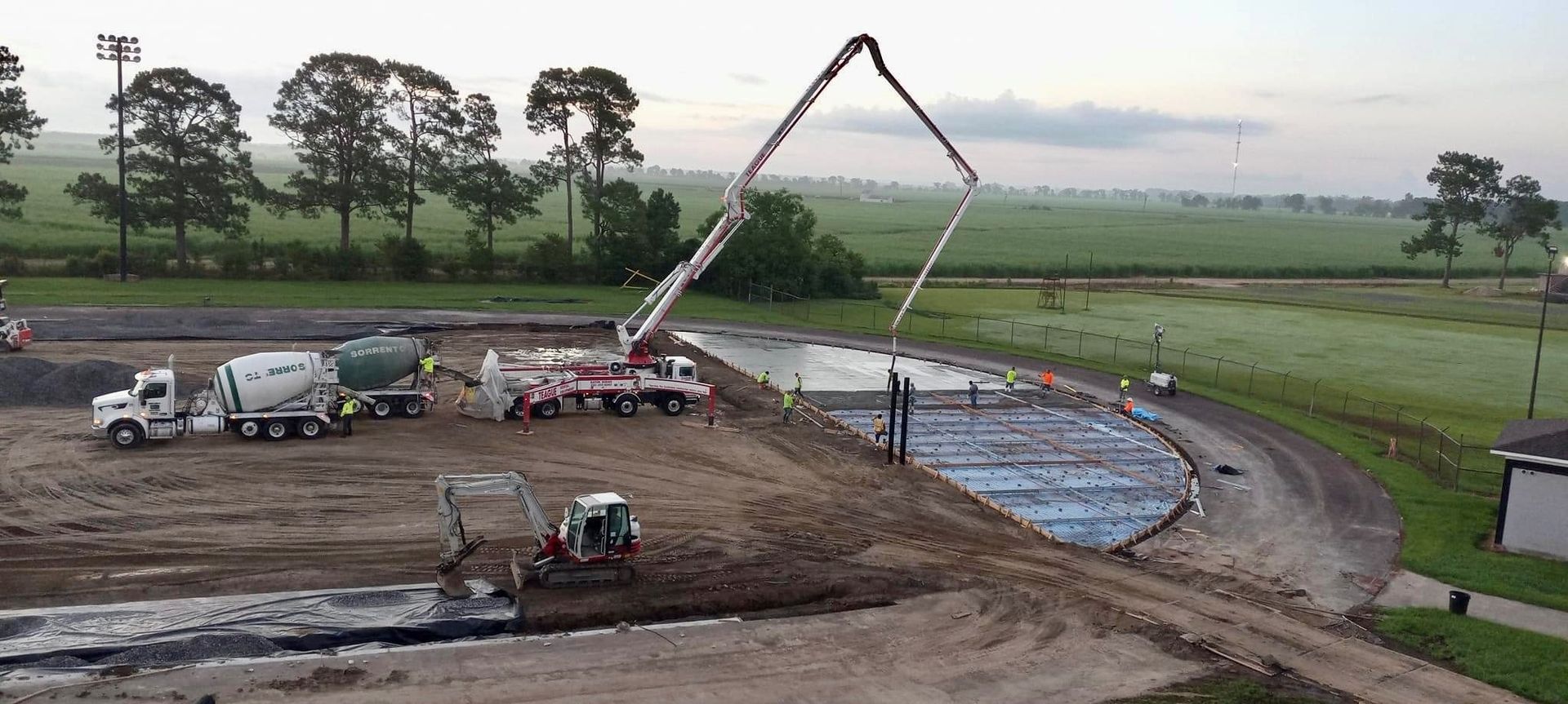 Construction site, concrete being poured into a circular foundation with trucks and machinery, green field in background.