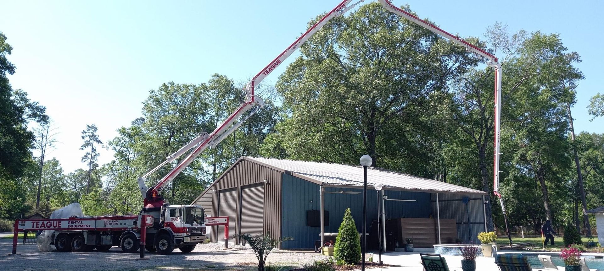 A concrete pump truck pours concrete on top of a blue and white building in a tree-filled area.