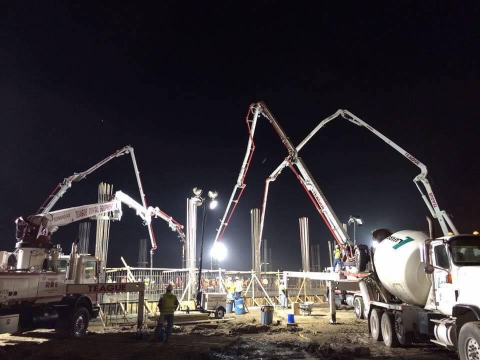 Construction site at night with concrete pumps pouring concrete into pillars. Workers are present, lit by floodlights.