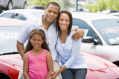 Family of three smiling near red car in a car lot.