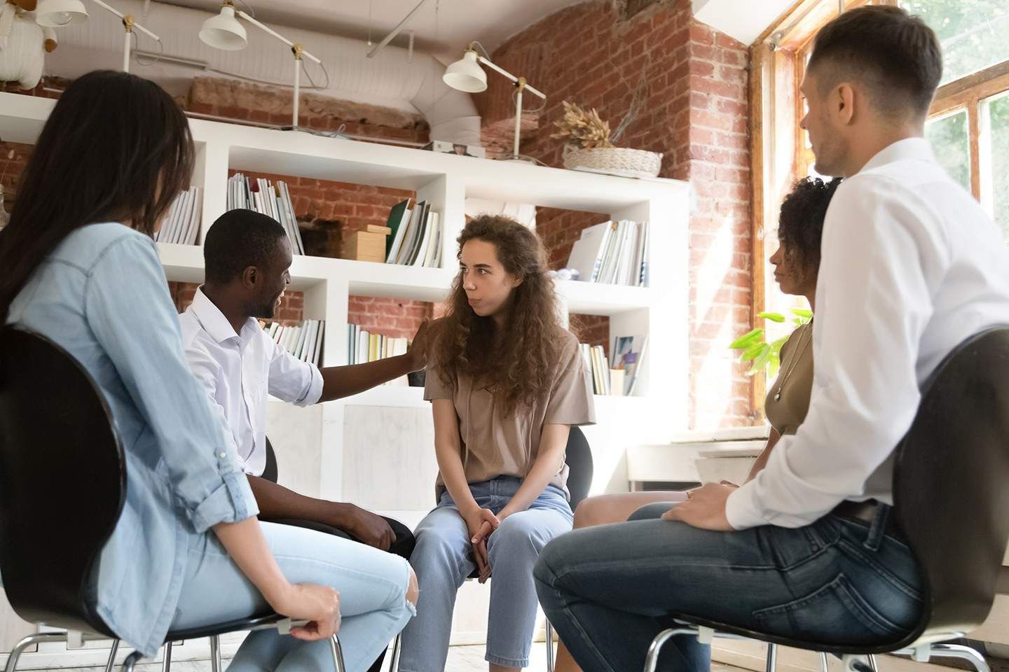 Group therapy session: People seated in a circle, offering support. Setting: Bright room with bookshelves and a window.
