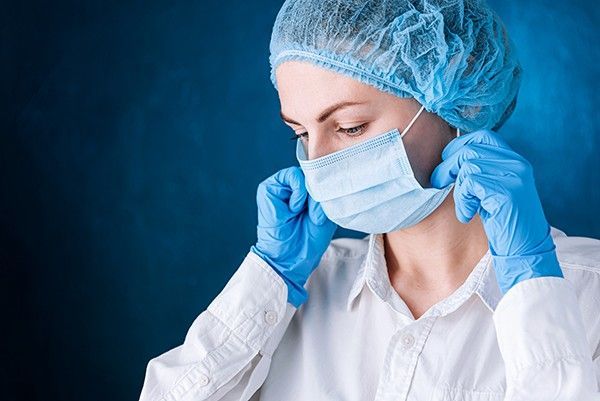 Person in blue gloves, cap, and mask, adjusting mask in a medical setting.