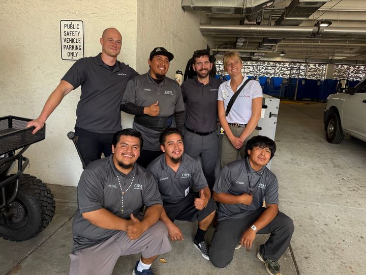 Group of six people posing in a garage beside a race car, wearing matching gray shirts