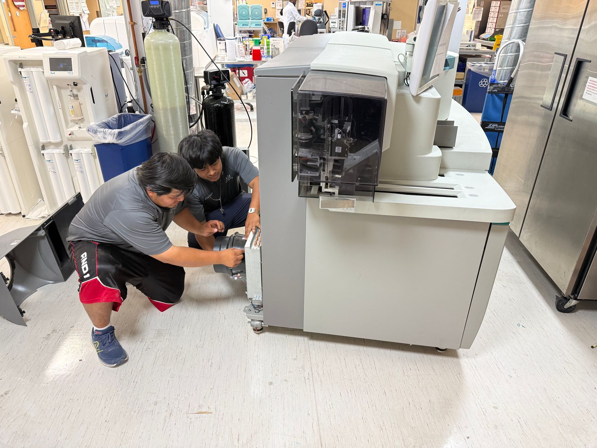 Two workers crouch beside a large industrial machine in a factory aisle.