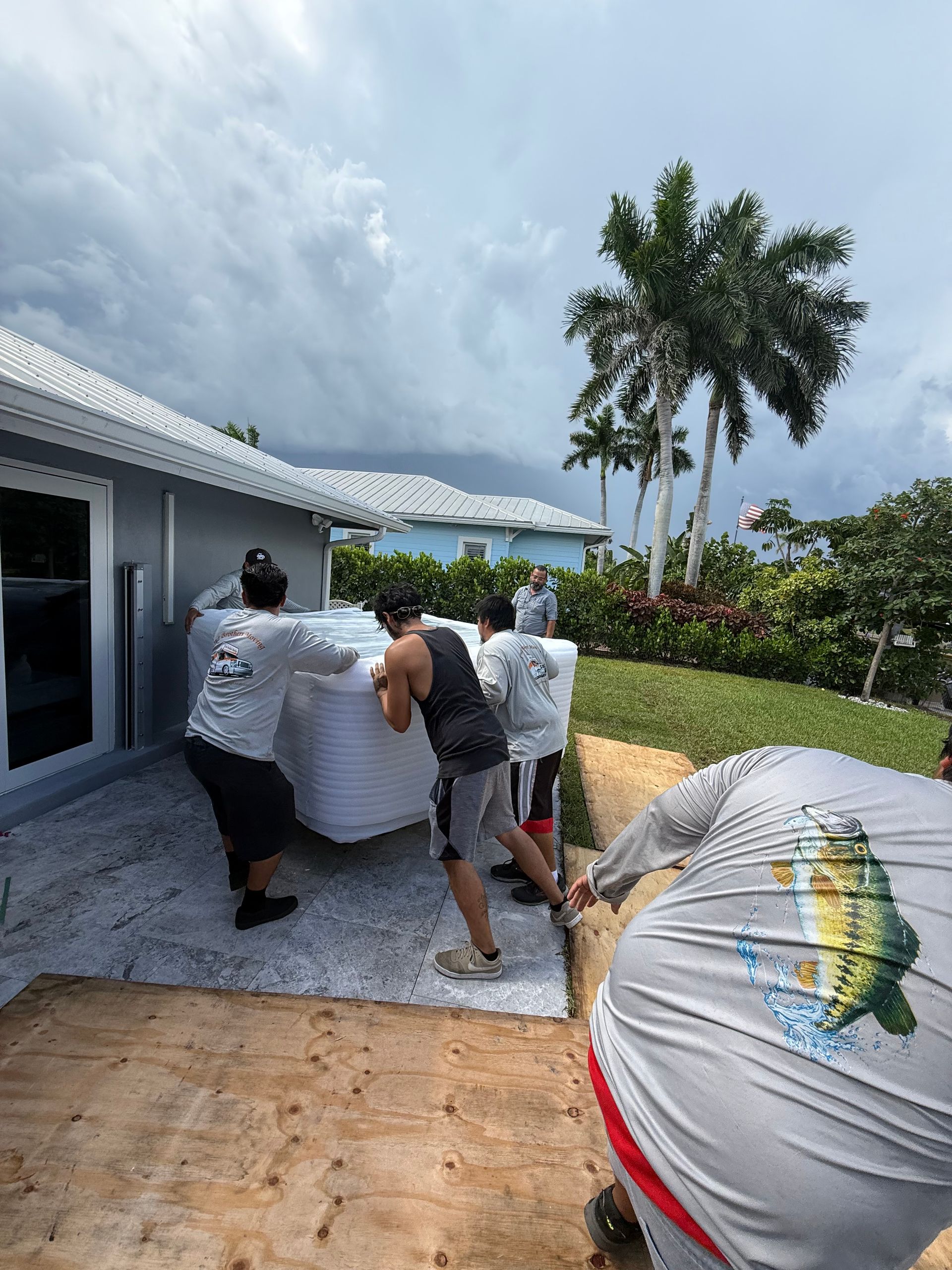 Three workers carry a wrapped item along a patio beside a building, under cloudy skies, with palm trees nearby.
