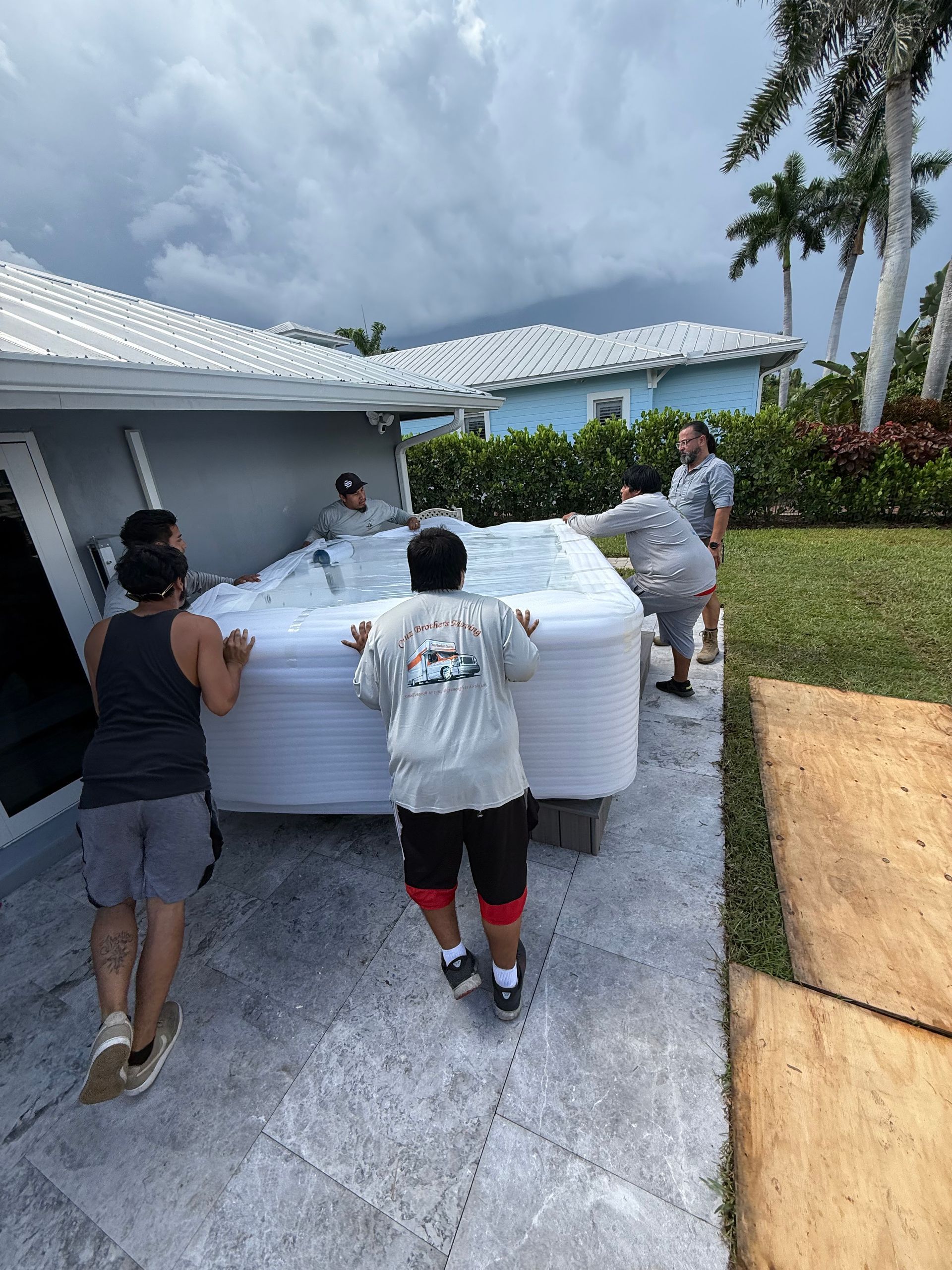 People moving a large white mattress across a driveway beside a house under cloudy skies