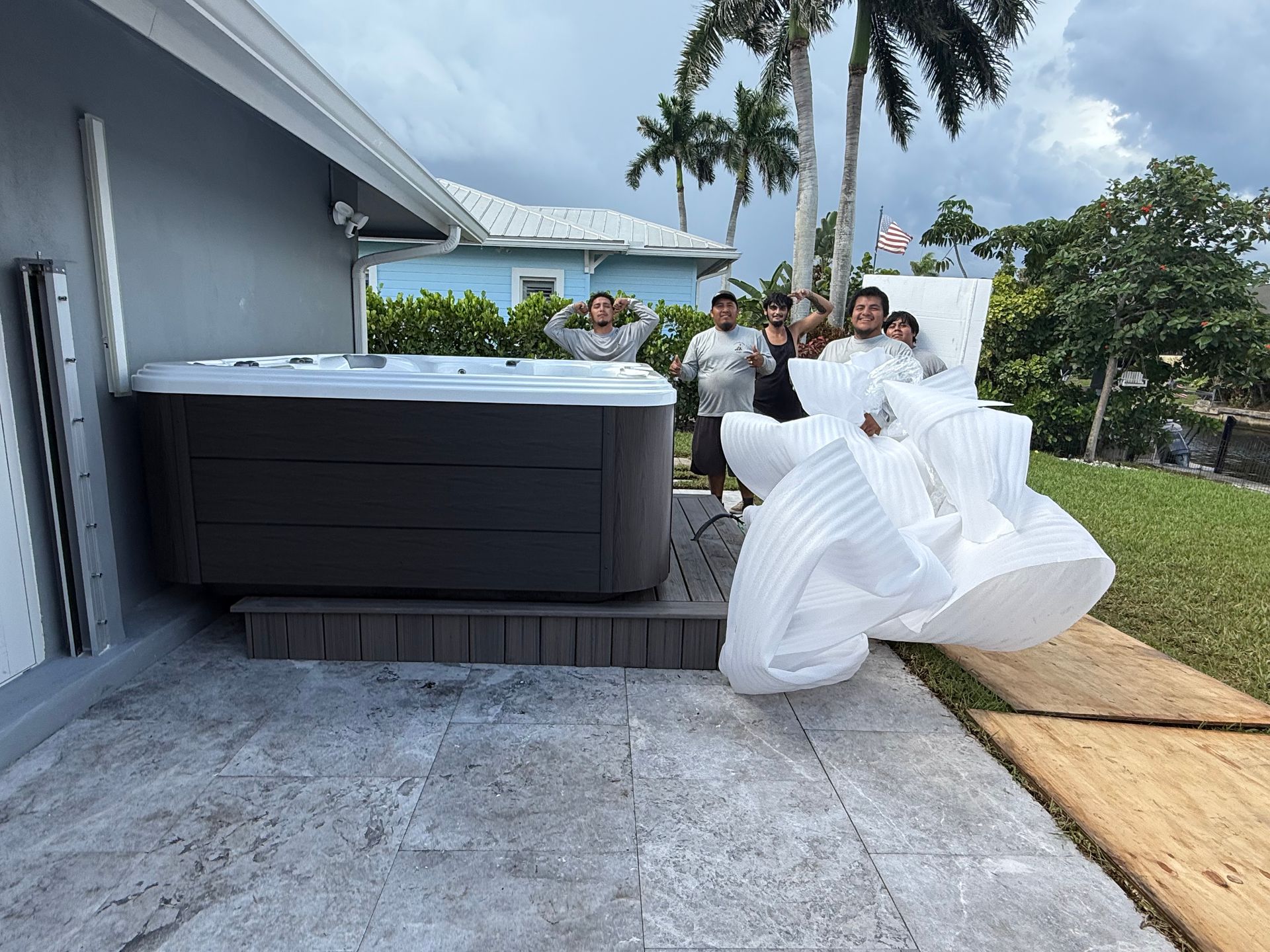 People carrying a white hot tub down a driveway beside a house, with a black hot tub nearby.
