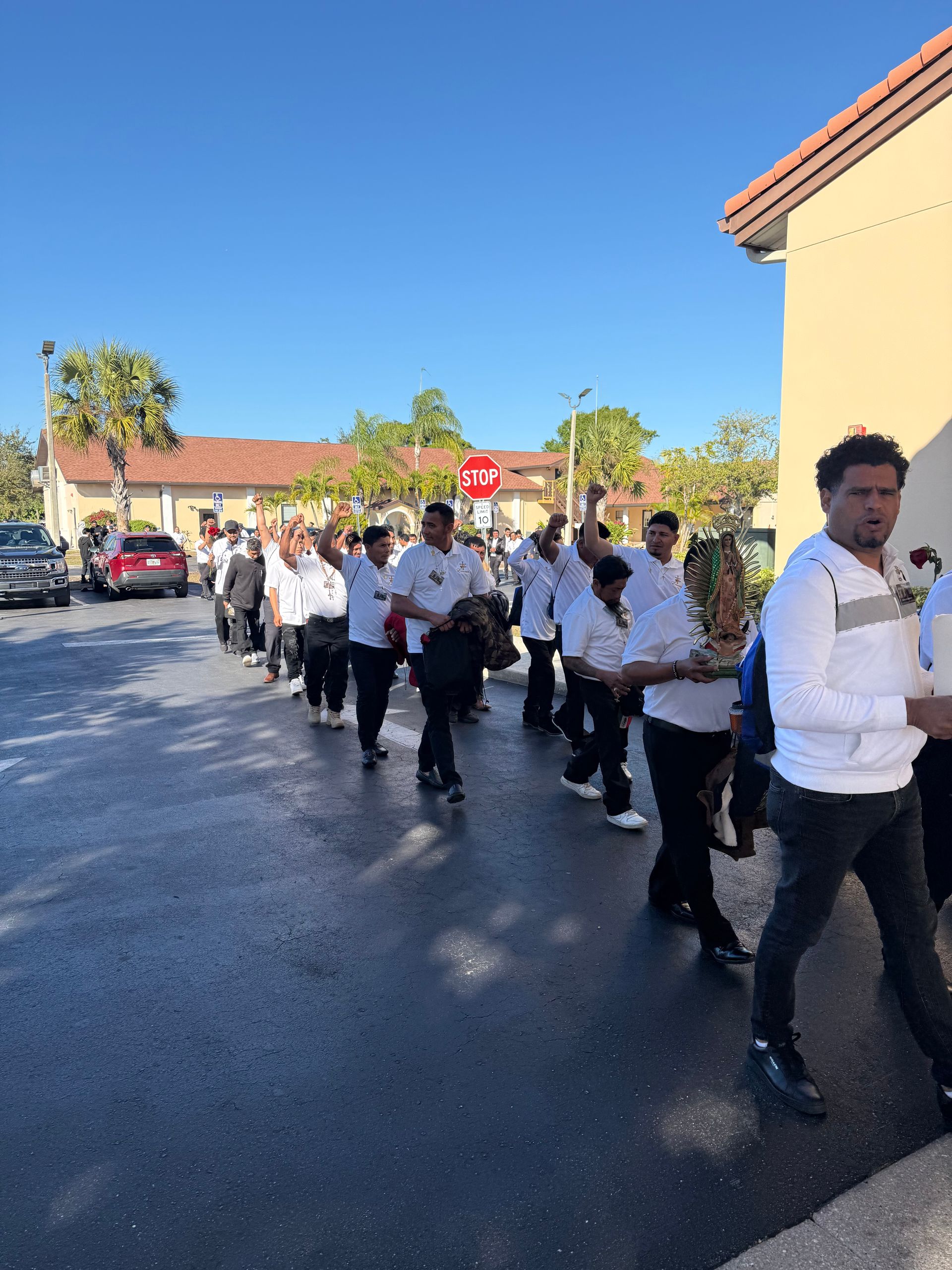 People in white shirts and black pants march in a line outdoors on a sunny street.