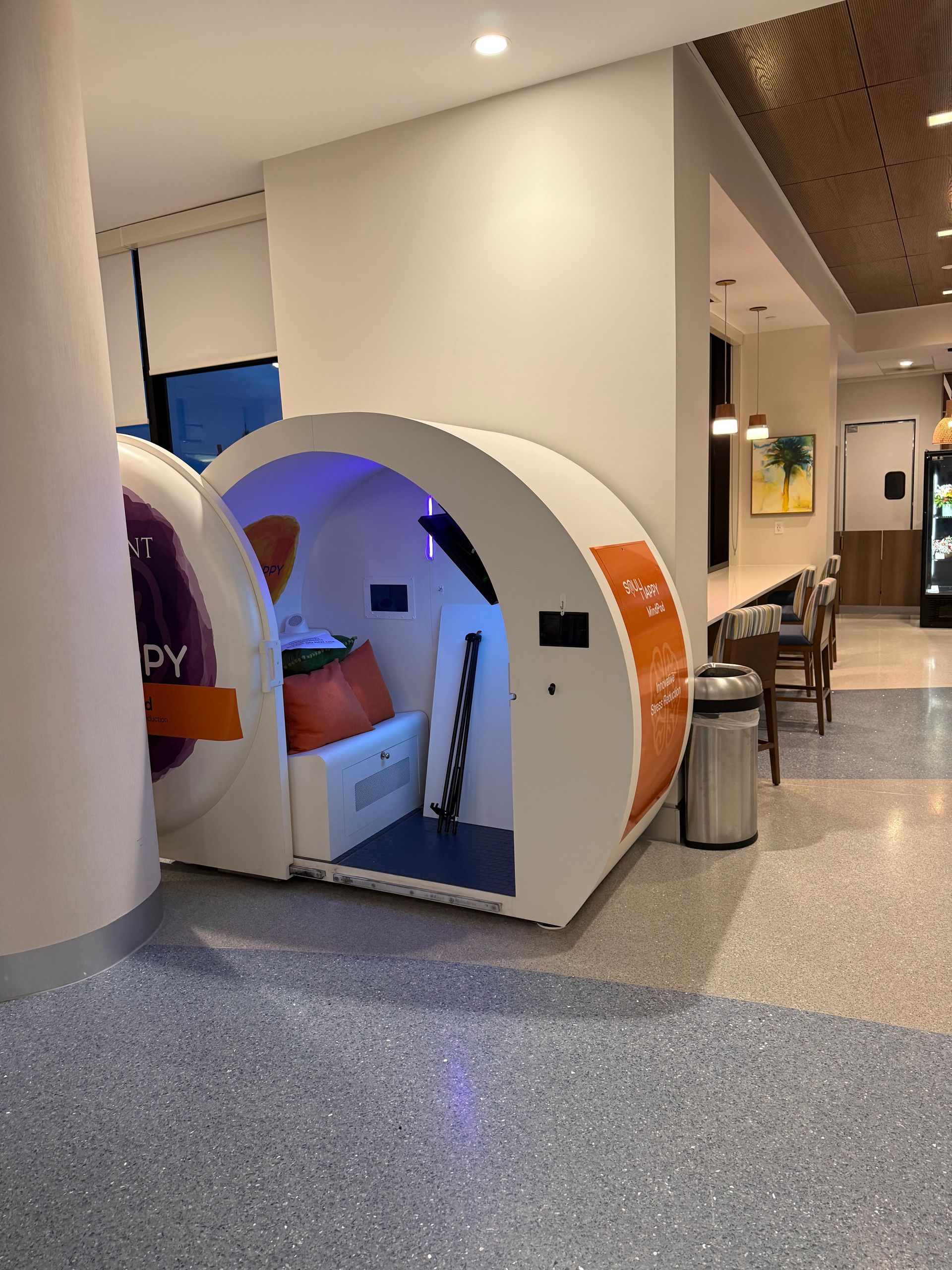 Pod-like booth in a modern hallway, lit in blue and orange, with a seated person inside.