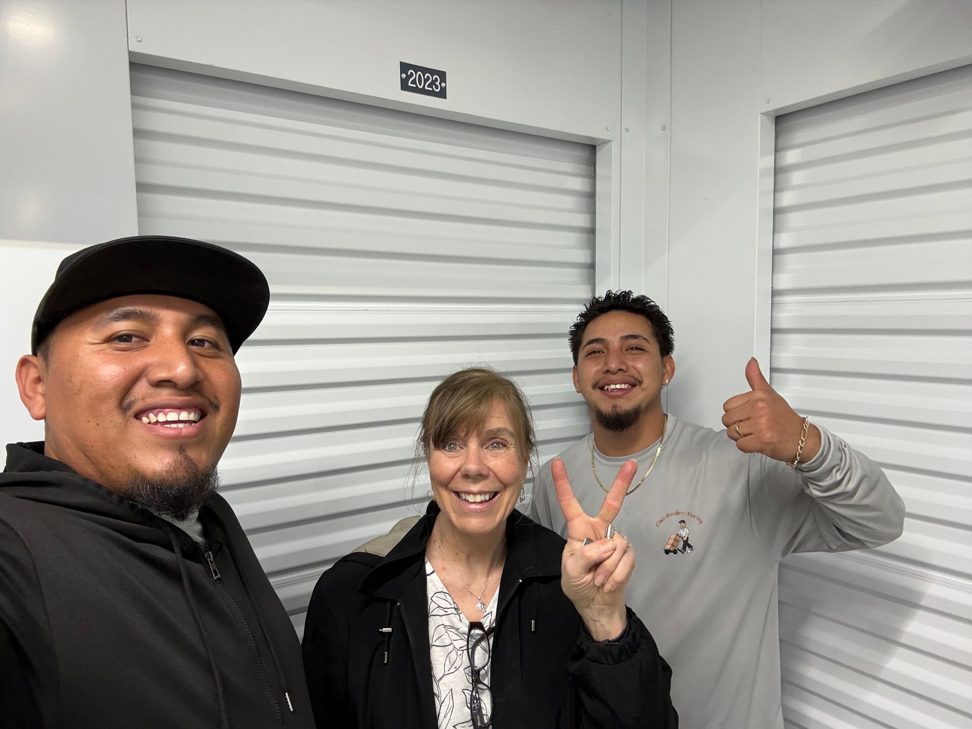 Three people smiling indoors, posing in front of a white metal roll-up door, one giving a thumbs-up.
