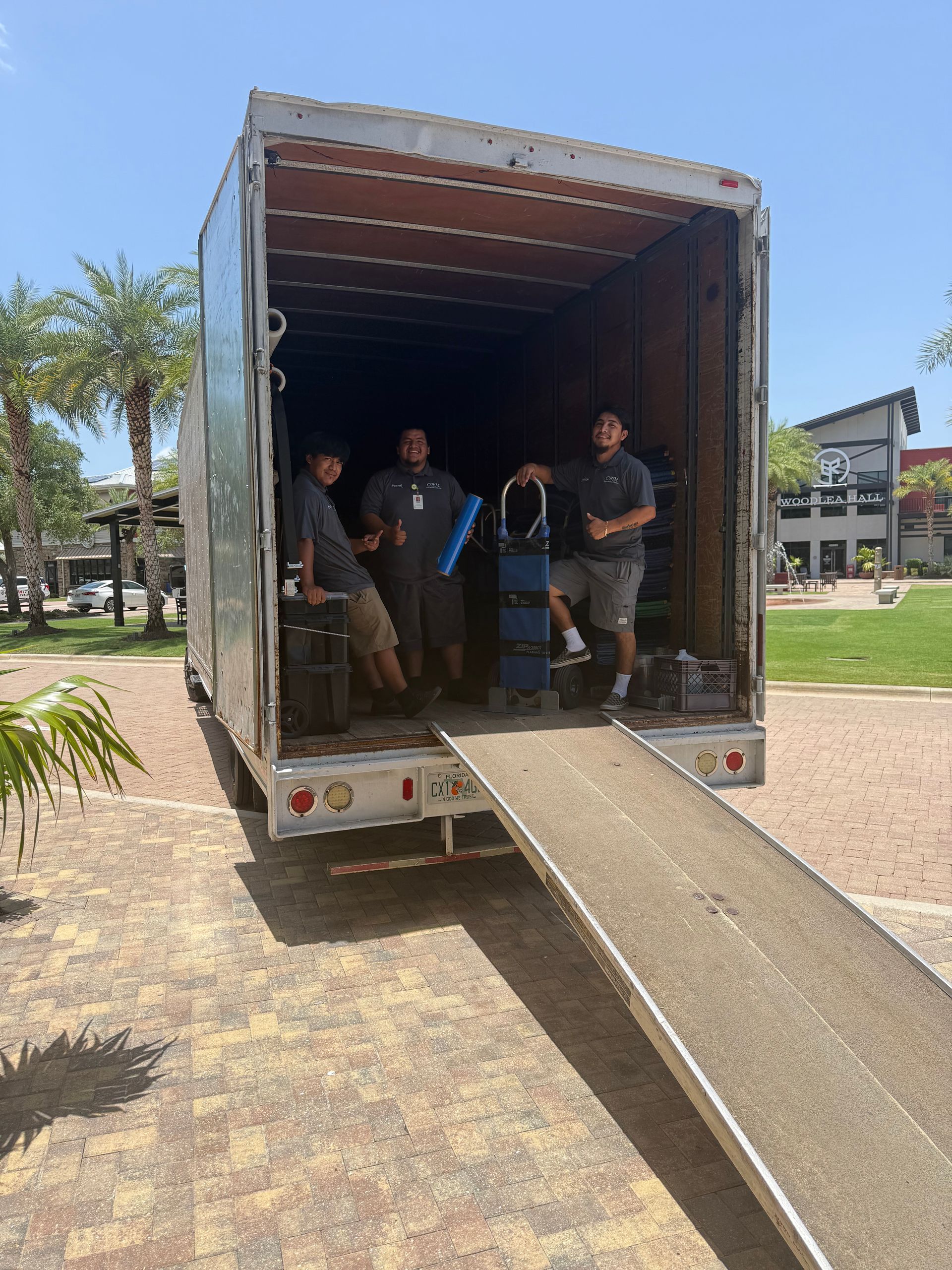 Truck parked with open cargo bay and ramp; three people stand inside in a sunny outdoor area.