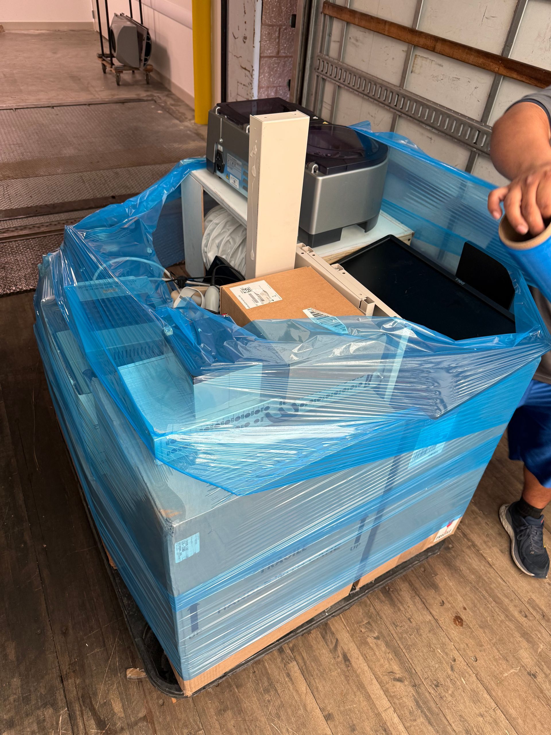 Blue-wrapped pallet with cardboard boxes beside a conveyor or packing machine in a warehouse